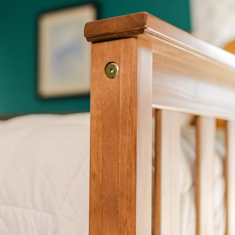 Close-up of solid wood bed frame headboard post with brass screw and cap, honey-stained finish, quilted bedding and teal wall background
