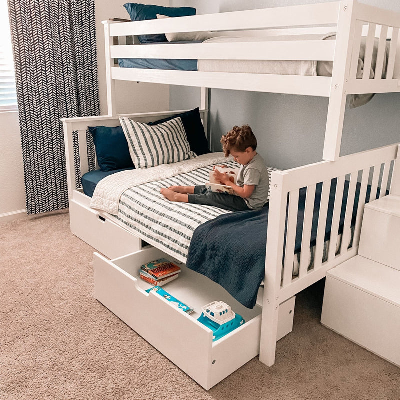 Space-saving white wooden bunk bed with under-bed storage drawer and navy-striped bedding, young child reading on the lower bed in a modern kids' bedroom