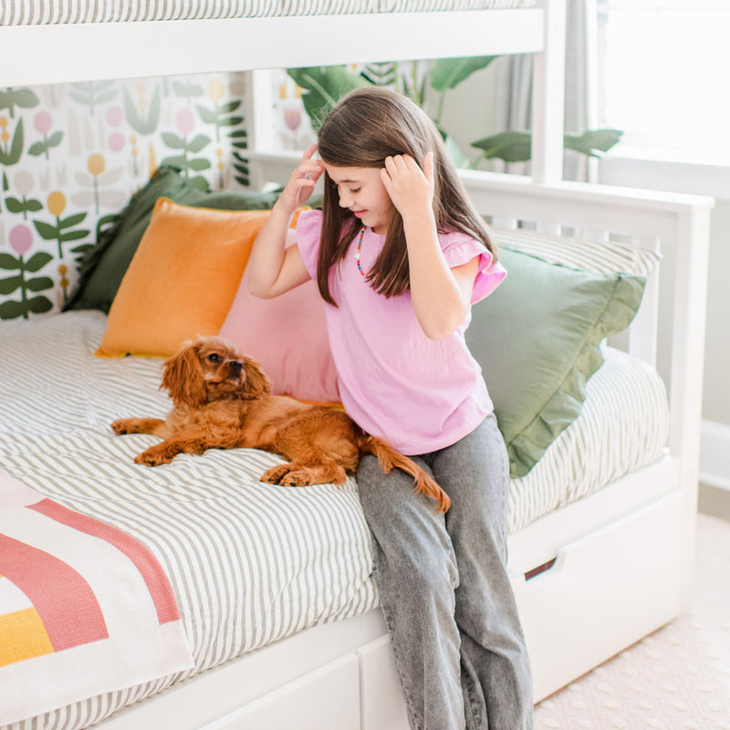 Young girl sitting on a striped bed beside a small brown dog in a brightly decorated kids' bedroom with floral wallpaper and pastel pillows