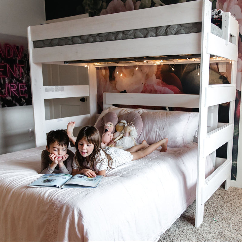Two children reading a picture book on a white bunk bed in a cozy pink bedroom with fairy lights, plush dolls and soft bedding