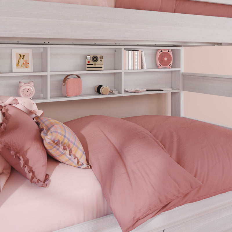Blush pink bunk bed with rumpled duvet and ruffled pillows, white built-in shelves displaying books, a pink alarm clock, retro instant camera and portable speaker