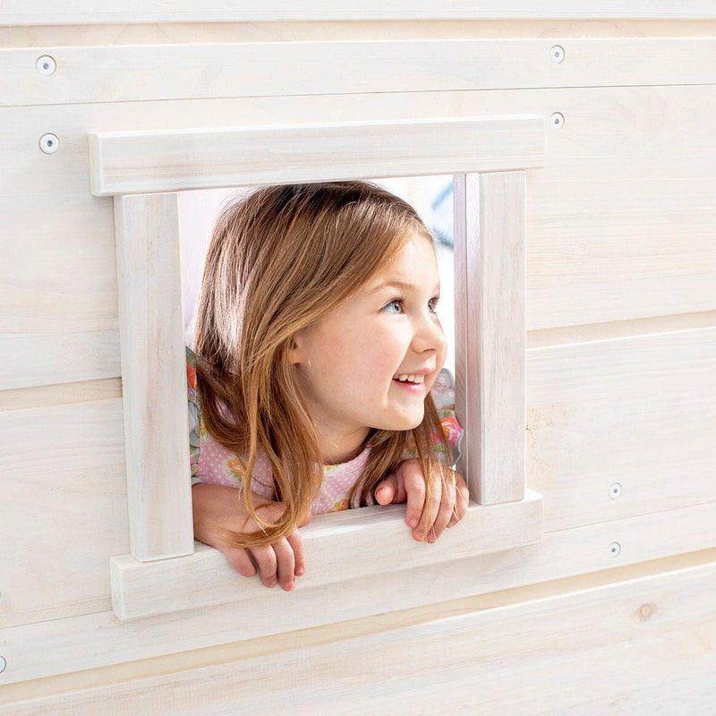 Smiling young girl peeking through the window of a whitewashed wooden children's playhouse, natural wood indoor playhouse toy for kids