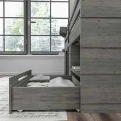 Gray wooden bunk bed with pull-out underbed storage drawer on caster wheels filled with folded linens in a bright modern bedroom by a large window