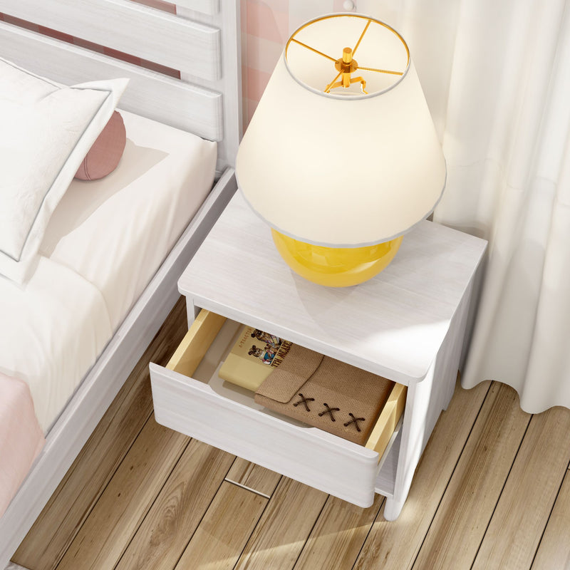 White nightstand beside white wooden bed with yellow ceramic table lamp and open drawer revealing a book and folded linen on hardwood floor