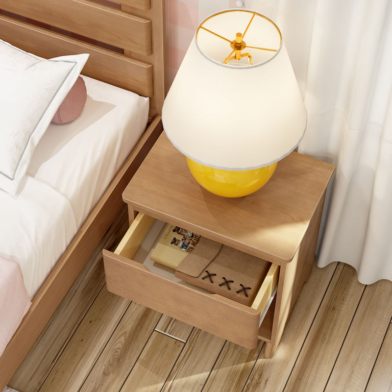Modern wooden bedside table with yellow lamp and open drawer holding a book and notebook next to a bed with white bedding on hardwood floor