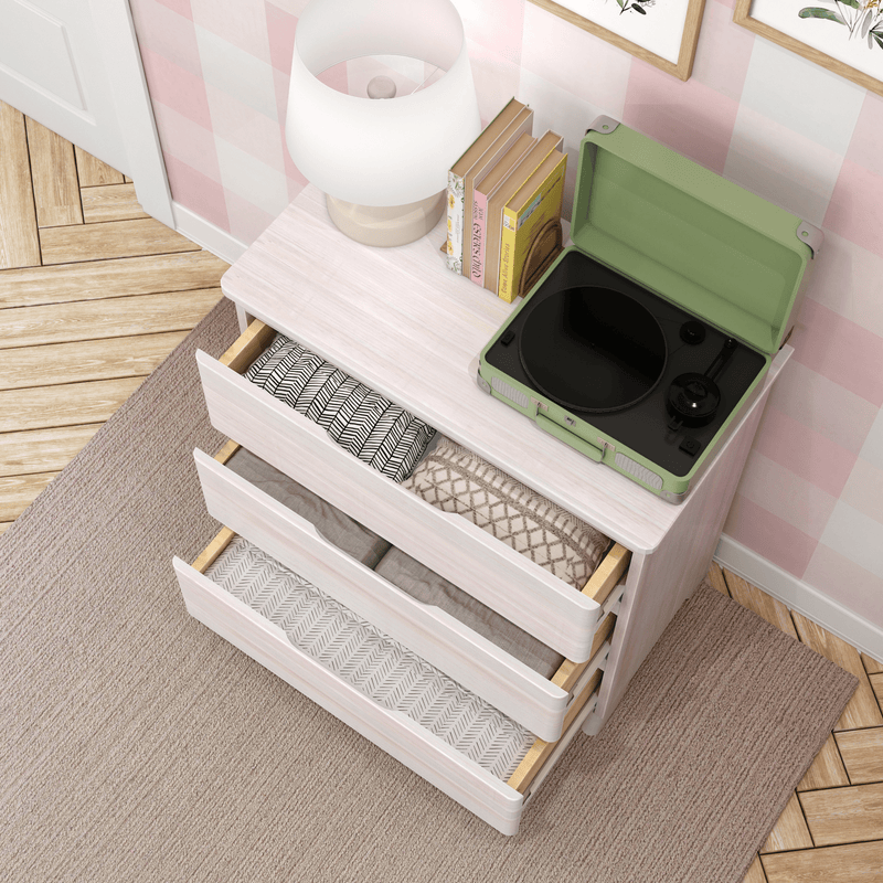 Top-down view of pastel pink dresser with open drawers of folded blankets, green portable record player, lamp and books in a nursery room