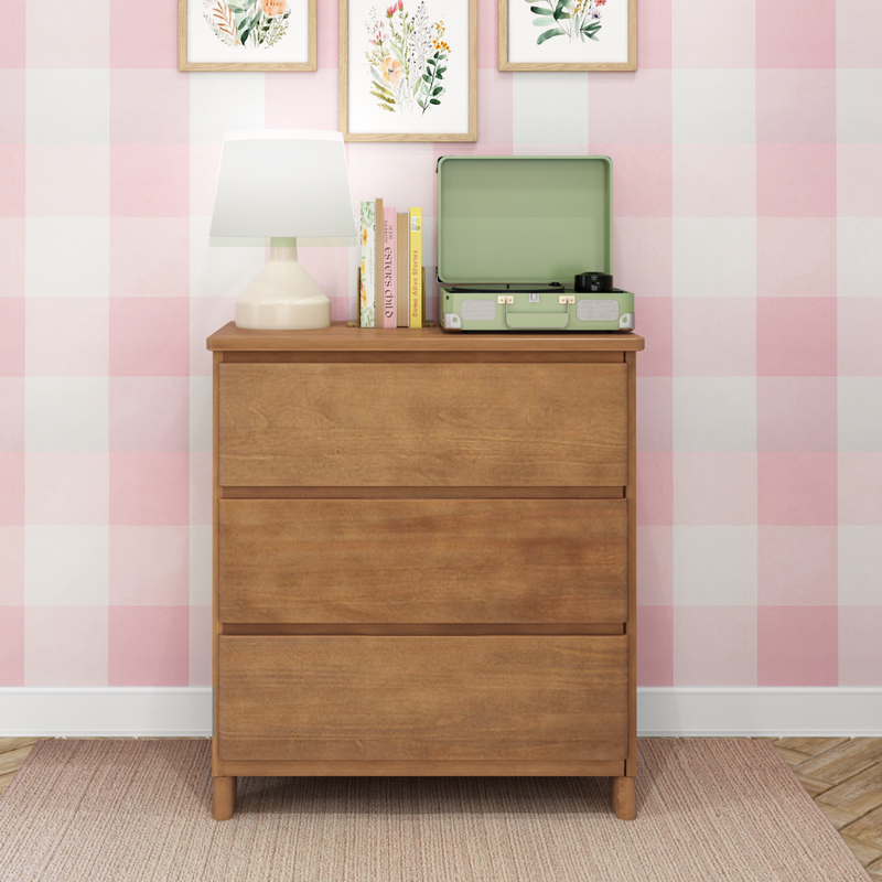 Wooden dresser topped with a white table lamp, stacked books and a green suitcase-style record player beneath framed botanical prints on pink gingham wallpaper