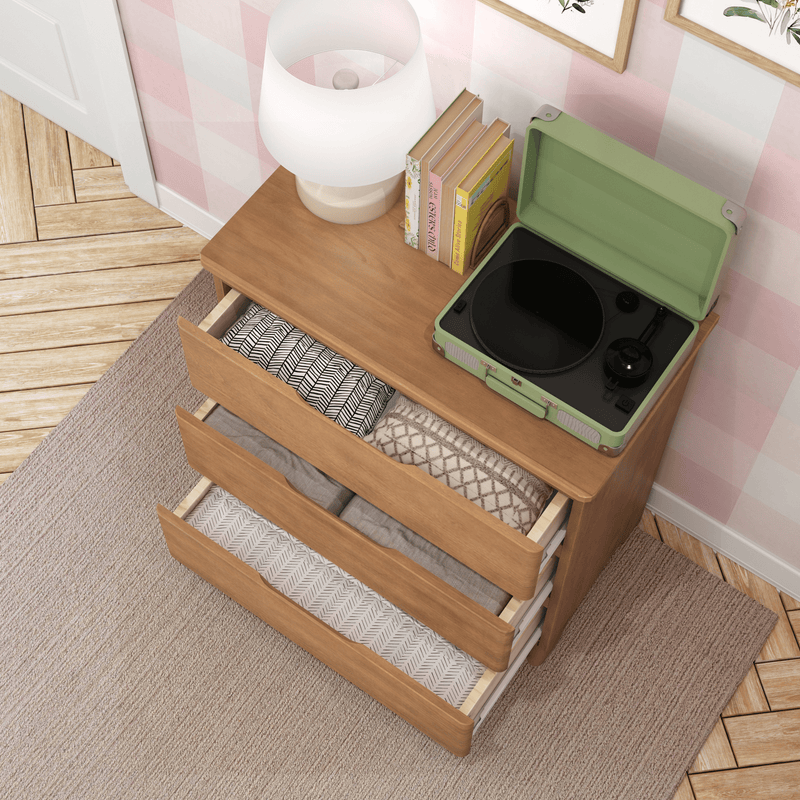 Wooden dresser with three open drawers of neatly folded linens, green portable vinyl record player, white table lamp and books on top against a pink checkered wall — bedroom storage and retro decor