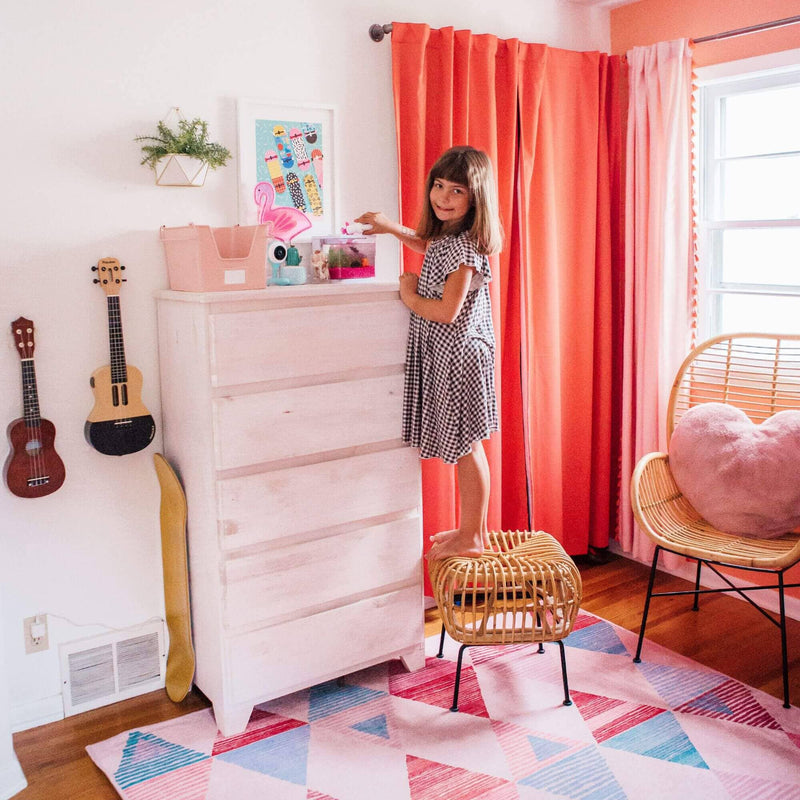 Kids' bedroom with coral curtains, white dresser, rattan chairs, geometric rug and a girl on a stool reaching for a toy