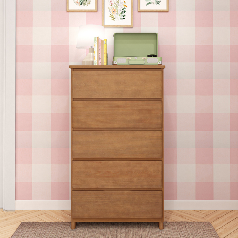 Wooden chest of drawers against pink gingham wallpaper in a nursery, topped with a table lamp, stacked books and a vintage green portable record player — kids room decor