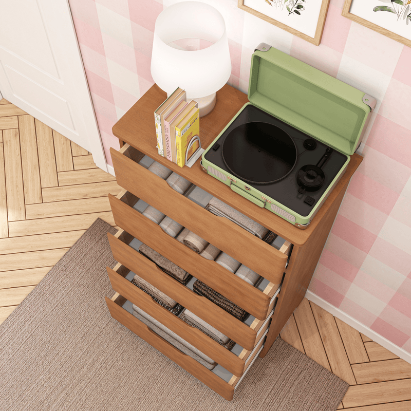Wooden dresser with open drawers displaying folded clothing, featuring a green portable record player and a lamp with books on top, set against a pink patterned wall.