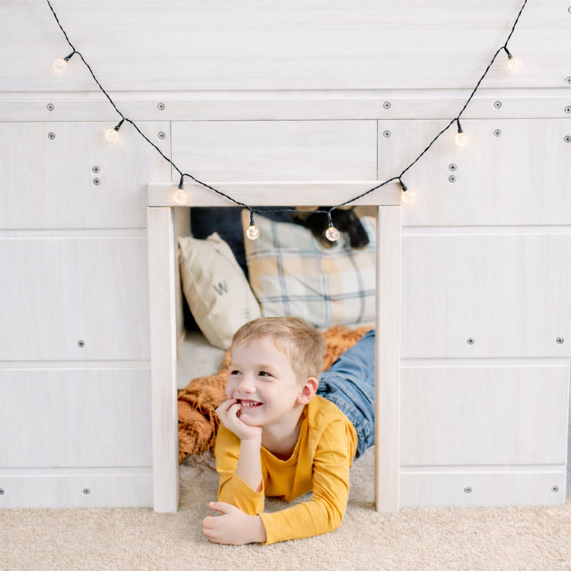 Smiling young boy in yellow shirt inside a cozy kids' indoor playhouse fort with string lights, plaid pillow and soft blanket