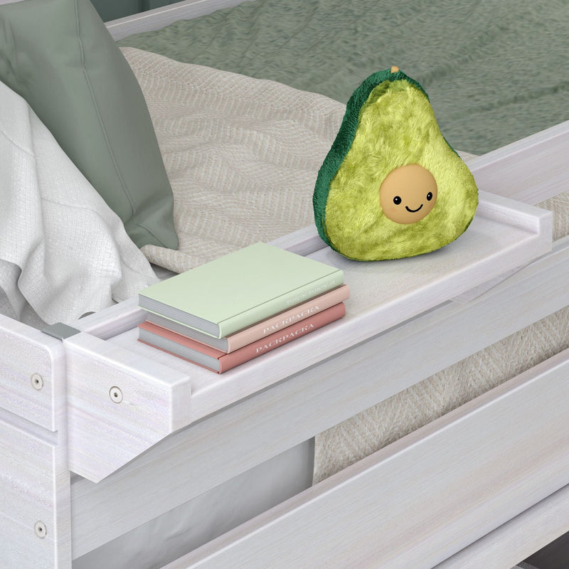 Plush smiling avocado pillow on a white wooden bed tray beside stacked pastel notebooks in a neutral kids’ bedroom