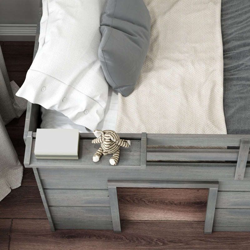 Top-down view of a gray wooden toddler bed in a neutral nursery with white and gray pillows, cream herringbone blanket, striped plush toy and books on hardwood floor