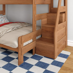 Solid wood twin-over-twin bunk bed with built-in storage staircase, beige bedding and car-print pillow in a child's bedroom on a blue-and-white checkered rug