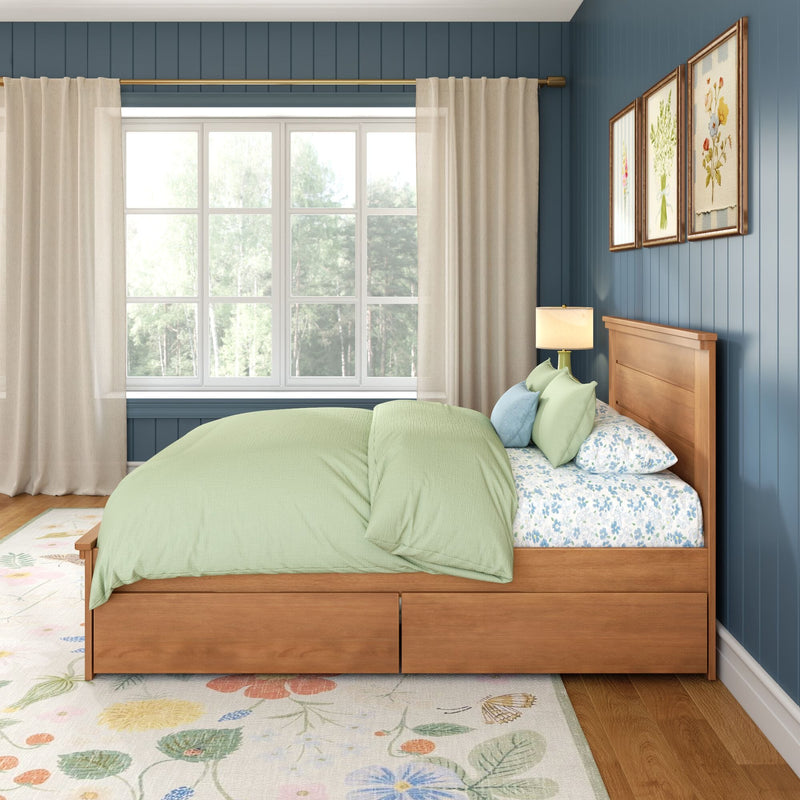 Bedroom with sage green bedding on wooden storage bed, floral sheets and rug, blue paneled wall, window with sheer curtains and brass curtain rod