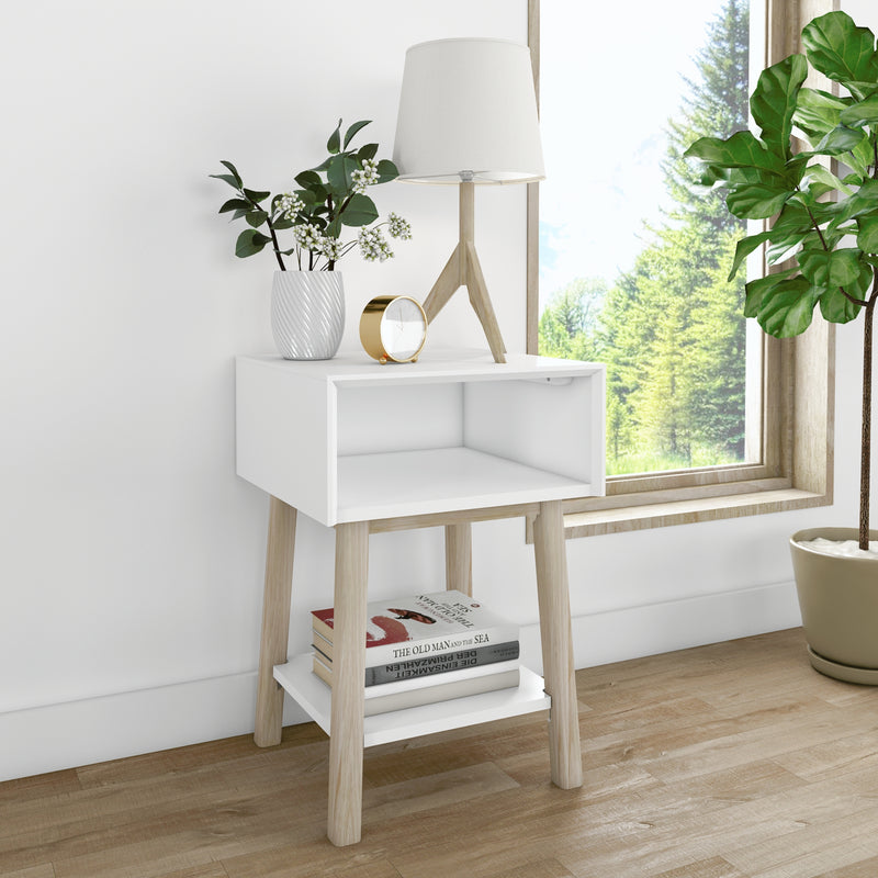 Modern minimalist white bedside table with wooden legs, lamp, potted plants and stacked books by a window overlooking green trees