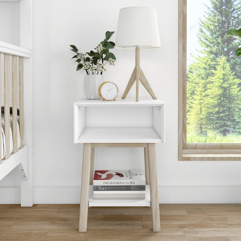 Scandinavian minimalist white nightstand with wooden legs, tripod table lamp, potted greenery, gold alarm clock and stacked books by a window showing a forest view