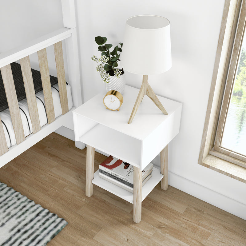 Modern white nightstand with light wood legs, tripod table lamp, gold alarm clock, potted plant and stacked books in a Scandinavian bedroom with hardwood floor and natural window light