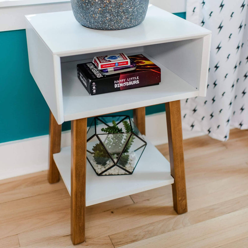 Modern white midcentury nightstand with wooden legs, geometric glass terrarium with succulents on lower shelf and Happy Little Dinosaurs game in cubby against teal accent wall