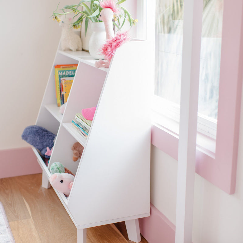 White angled kids bookshelf storage organizer with cubbies holding stuffed animals, picture books and plush toys in a pink-accented sunlit nursery on hardwood floors