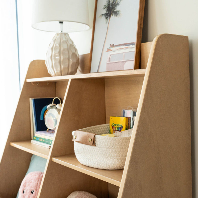 Wooden leaning bookshelf organizer for kids in nursery with ceramic lamp, framed palm print, woven storage basket holding Crayola crayons and children's books