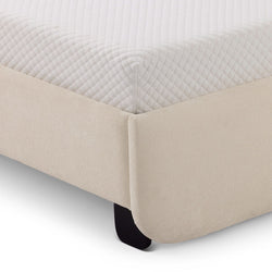 Corner view of a beige fabric bed frame with a white quilted mattress in a modern bedroom setting.
