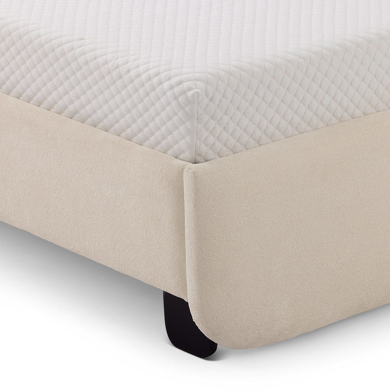 Corner view of a beige fabric bed frame with a white quilted mattress in a modern bedroom setting.