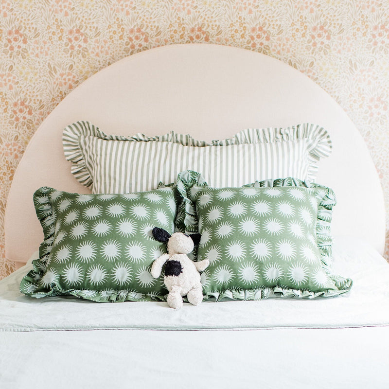 Nursery bed with round beige headboard, floral wallpaper backdrop, green sunburst-pattern pillows, striped ruffle pillow and plush toy