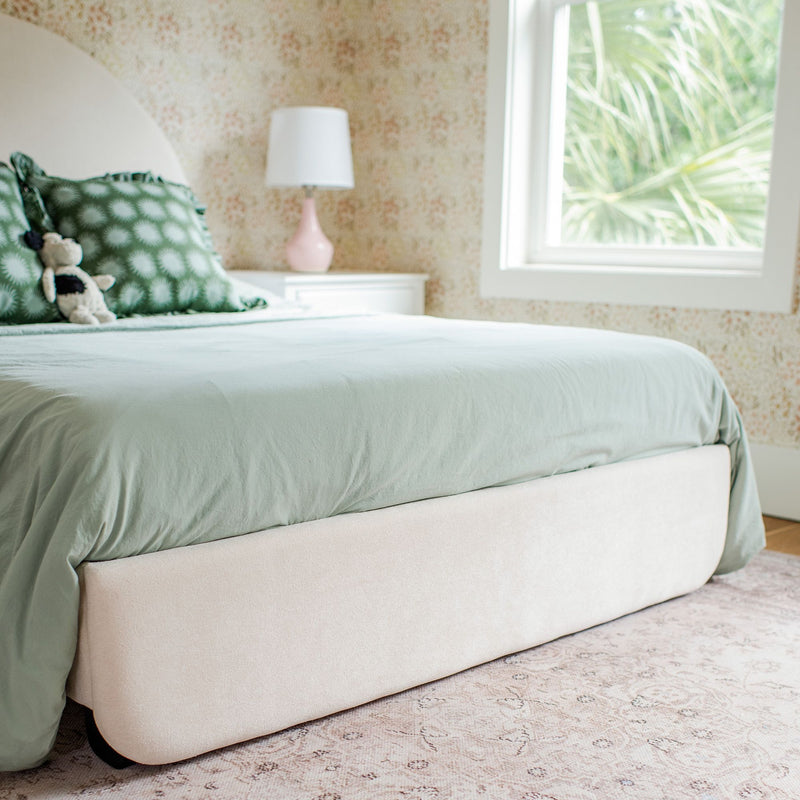 Sage green bedding on an upholstered bed with patterned green pillows, stuffed toy, pink bedside lamp and floral wallpaper by a sunlit window