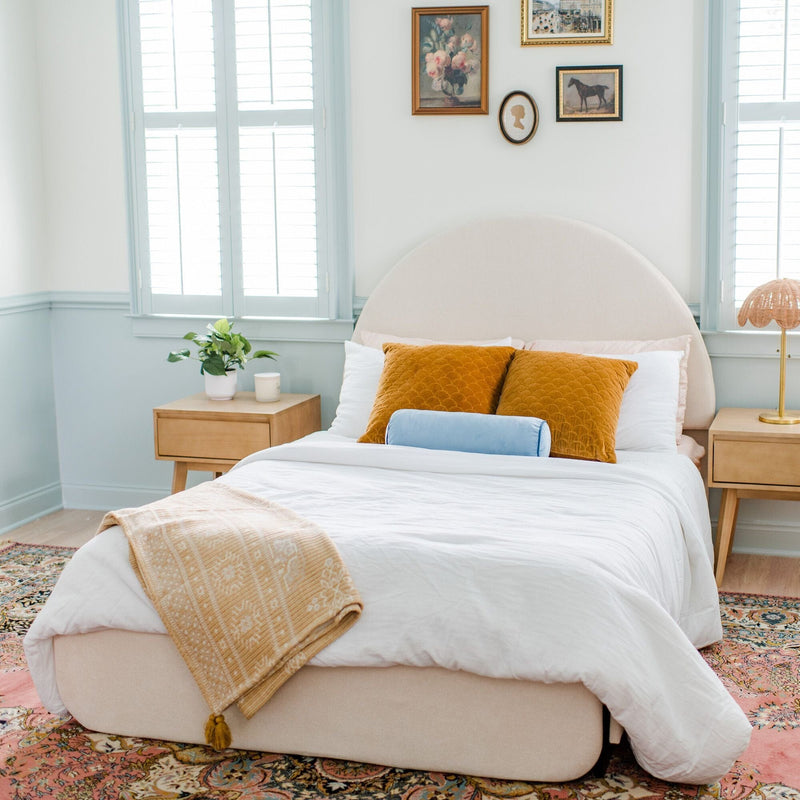 Bright cozy bedroom with rounded upholstered headboard, white bedding, mustard velvet pillows, light blue bolster, wooden nightstands, shuttered windows, potted plant, brass floor lamp and patterned area rug