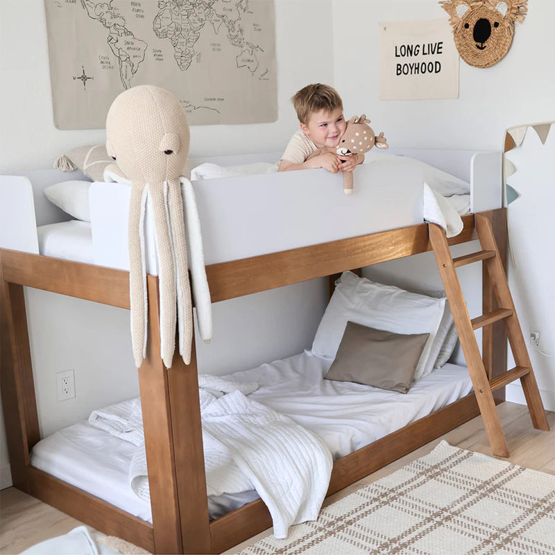 Neutral Scandinavian kids bunk bed with white linens, wooden frame, knitted octopus hanging, boy holding deer plush, map wall art and Long Live Boyhood sign