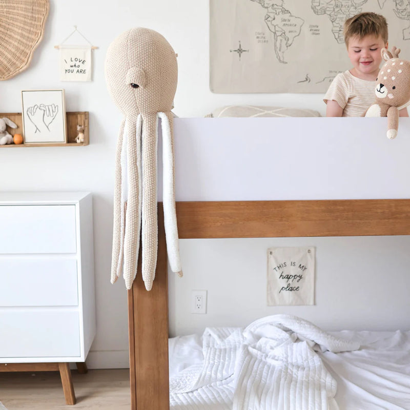 Minimalist neutral nursery decor: wooden bunk bed with white bedding, hanging crochet octopus toy, plush deer stuffed animal and world map wall art
