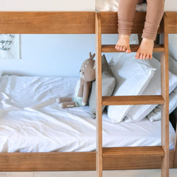Child's feet on a wooden bunk bed ladder with a plush toy on a white sheeted bed.