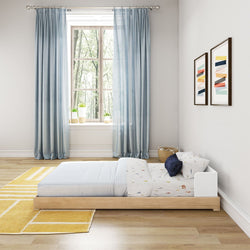 Minimalist kids bedroom with floor bed, blue curtains, yellow rug, and wall art in bright daylight.