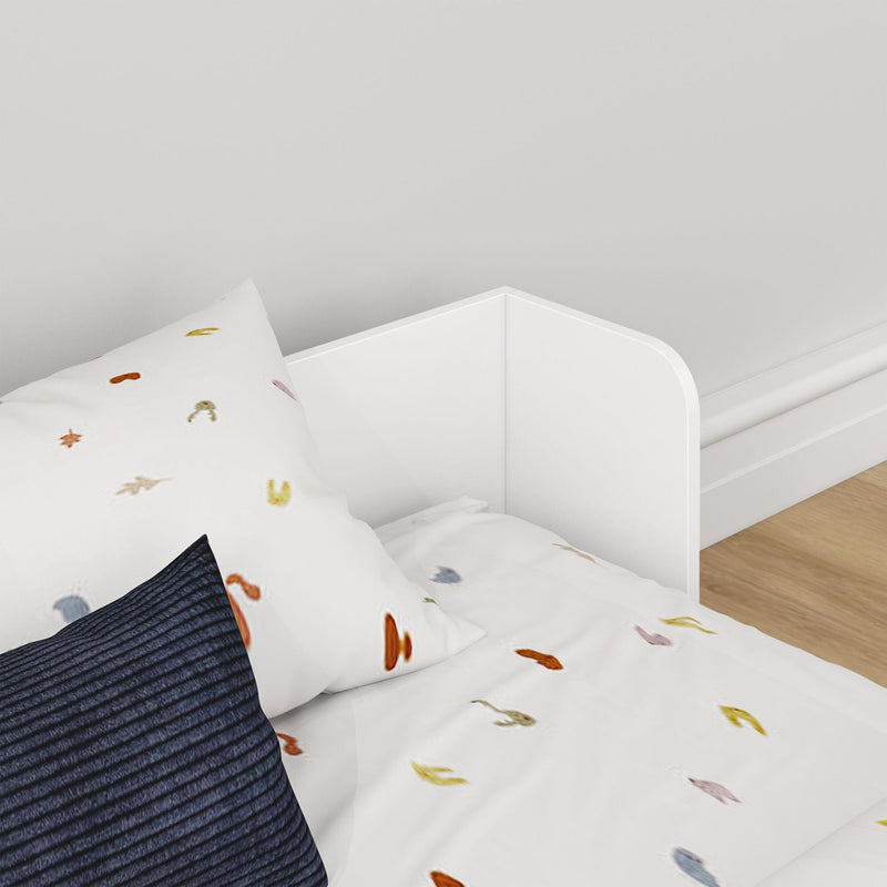 Close-up of a modern white wooden toddler bed and rail with leaf-print white duvet and navy textured pillow on a light wood nursery floor