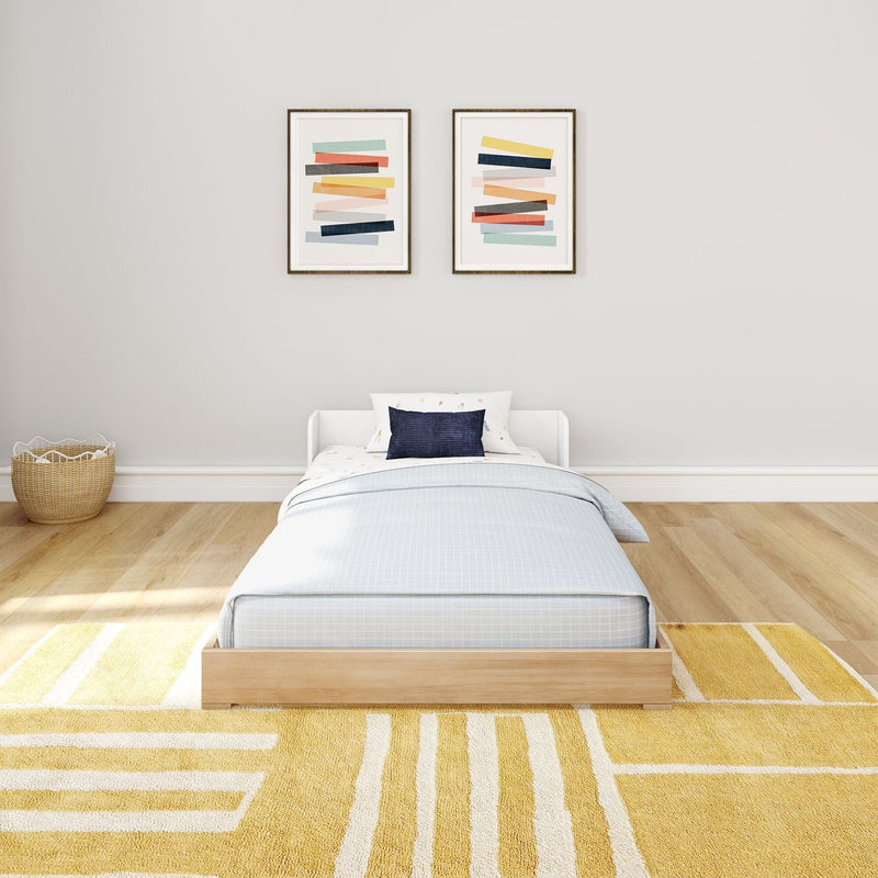Minimalist children's bedroom with light wood platform bed, blue pillow, two abstract framed prints, wicker basket and yellow geometric rug.