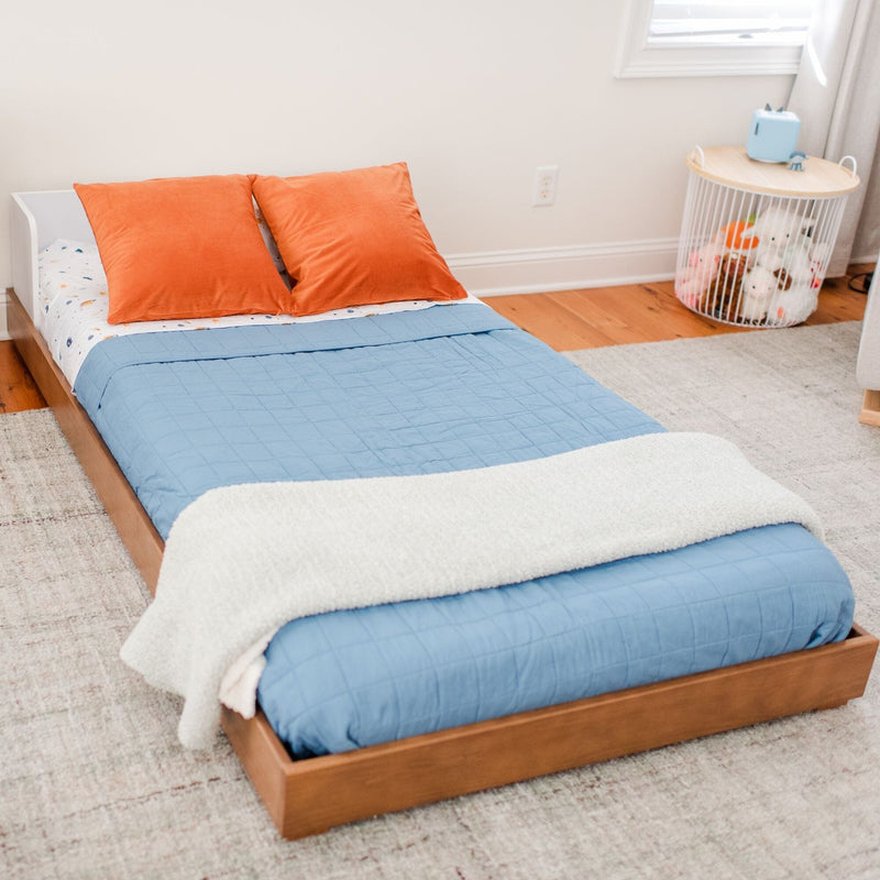 Low wooden toddler bed with blue quilt and orange pillows in bright kids bedroom, modern nursery decor with toy storage basket