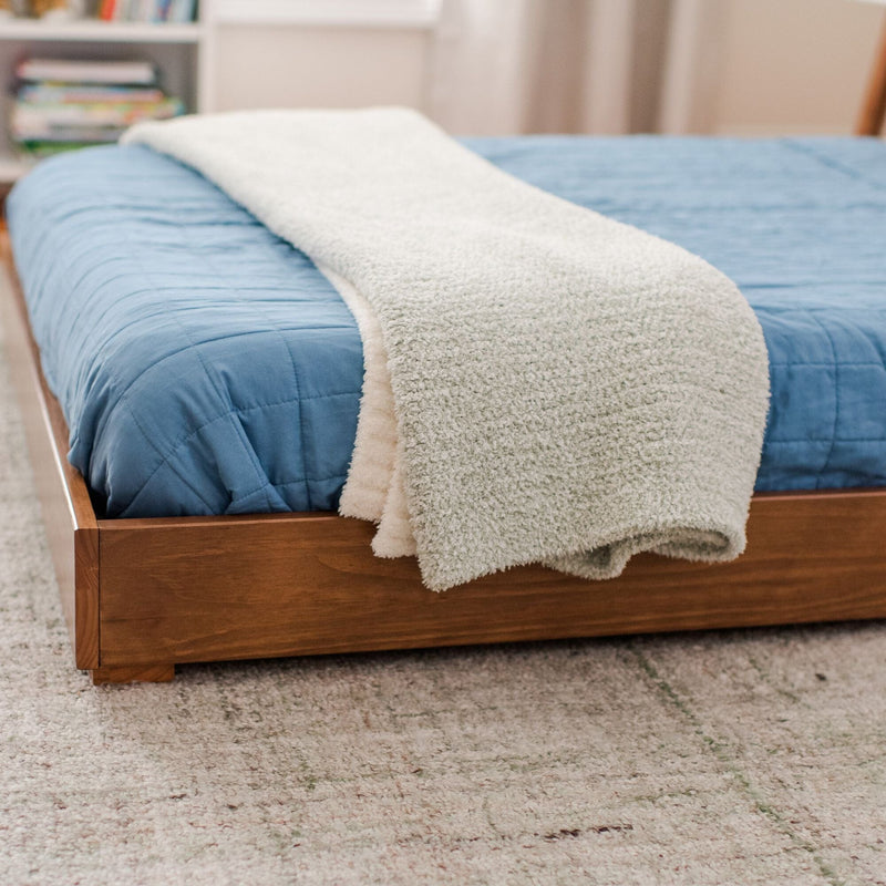 Cozy bedroom close-up of a wooden platform bed with a blue quilted comforter and cream sherpa throw blanket folded at the foot on a neutral textured rug, modern minimalist bedroom decor