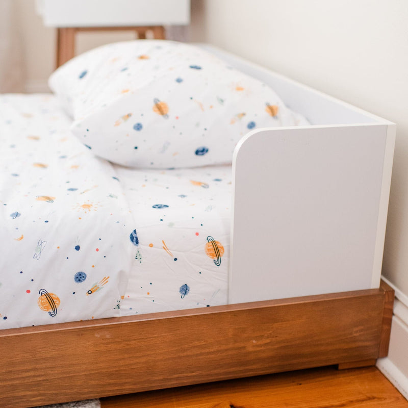 White toddler bed with wooden frame and safety rail, fitted with white space-themed planet print sheets and pillow — nursery bedding