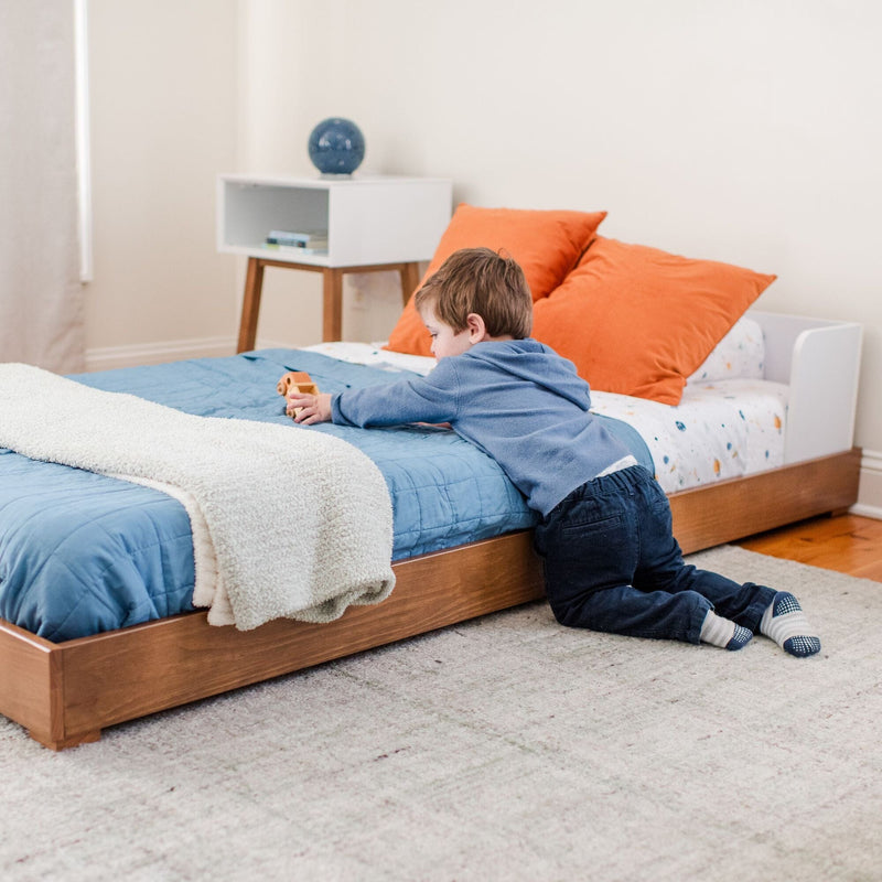 Child playing with wooden toy car on bed in modern kids bedroom with blue quilt, orange pillows and wooden nightstand