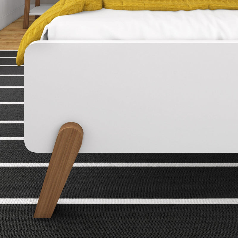 Modern bed with white frame, wooden legs, and yellow bedding on black striped rug.
