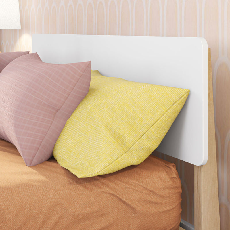 Close-up of pink and yellow decorative pillows on a bed with a modern white rounded headboard, peach textured bedding and wood frame against patterned wallpaper