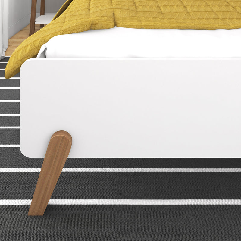 Close-up of modern white platform bed corner with mustard yellow quilt, angled wooden legs and black-and-white striped rug in minimalist bedroom interior