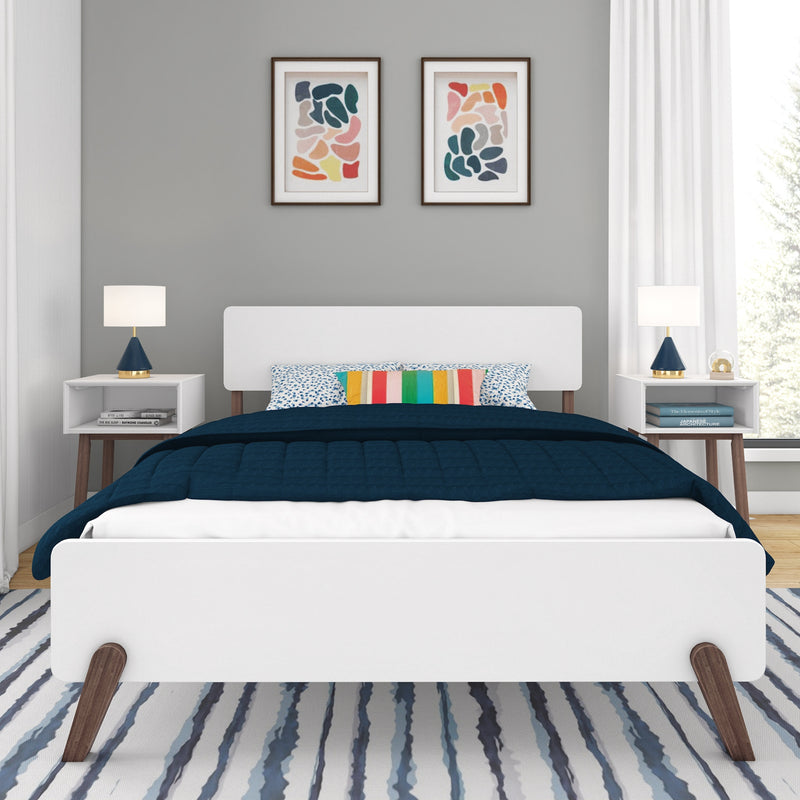 Contemporary bedroom with white platform bed, navy quilt, colorful striped accent pillow, blue table lamps on mid-century nightstands, gray wall with framed abstract art