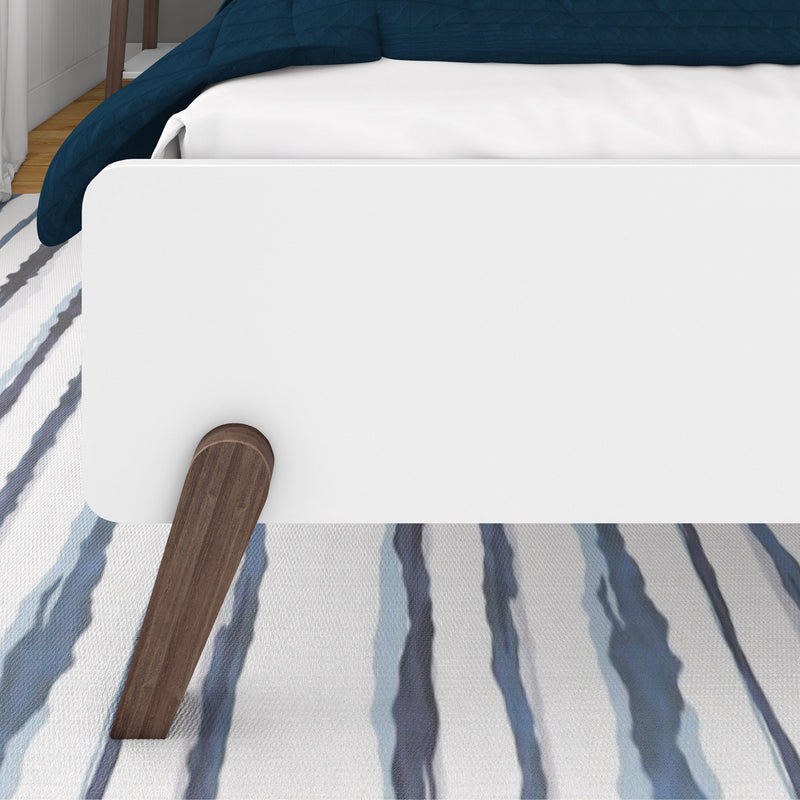 Modern white platform bed with navy blue quilt and white sheets, wooden legs on a blue-striped area rug in a bright contemporary bedroom
