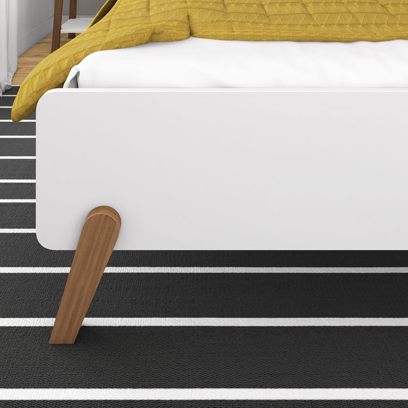 Close-up of modern white platform bed footboard with mustard yellow quilt, wooden bed leg and black-and-white striped rug in minimalist bedroom