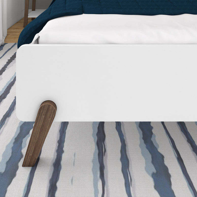 Close-up of white platform bed footboard with white mattress and navy quilt, wooden bed leg on a blue-striped area rug in a modern bedroom