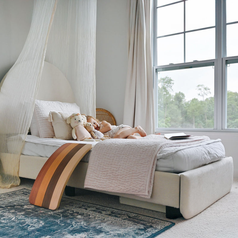 Cozy neutral nursery with canopy bed, soft pastel quilt, stuffed animals and wooden balance board beside a large window with natural light