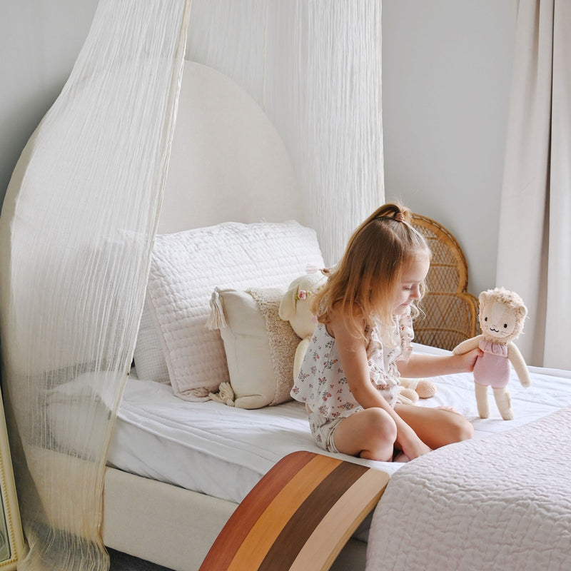Girl playing with a stuffed doll on a canopy bed in a neutral nursery with quilted bedding, wicker chair and wood rocker